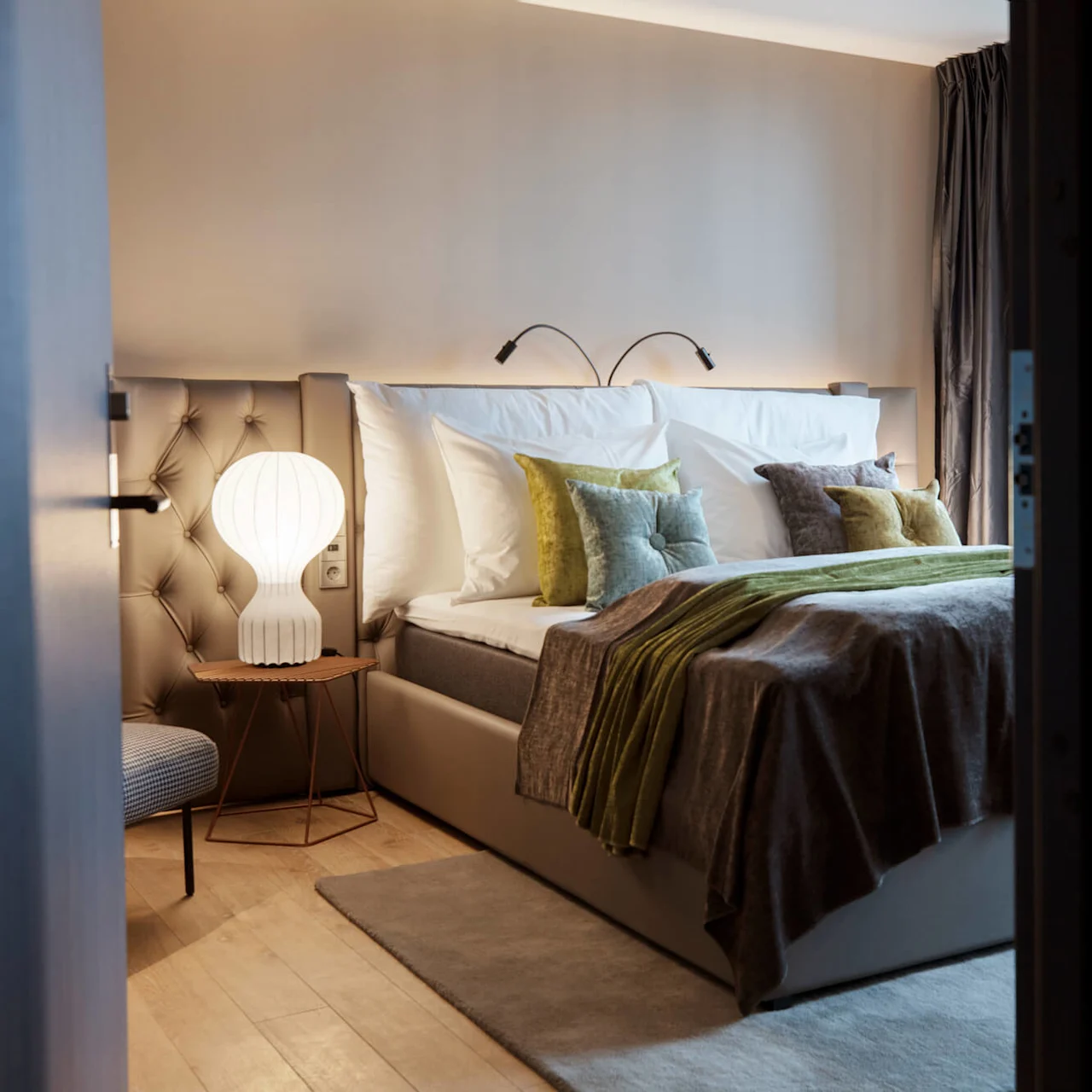 A cozy bedroom has a neatly made bed with white and green pillows and a gray blanket. A glowing lamp sits on a small table beside the bed in a softly lit room.