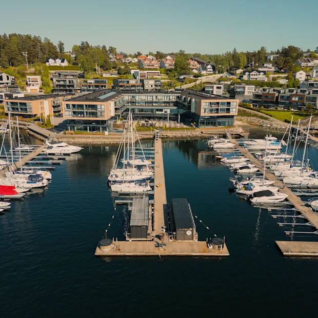 En luftfoto af en livlig marina med mange både fortøjet, mod en baggrund af moderne bygninger og huse på en bjergskråning.