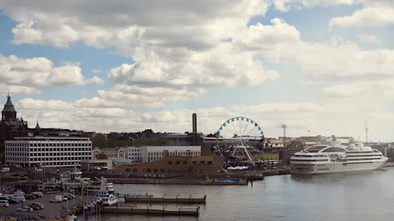Et stort pariserhjul står stille ved en havn, omgivet af bygninger og et fortøjet krydstogtskib under en delvis overskyet himmel.
