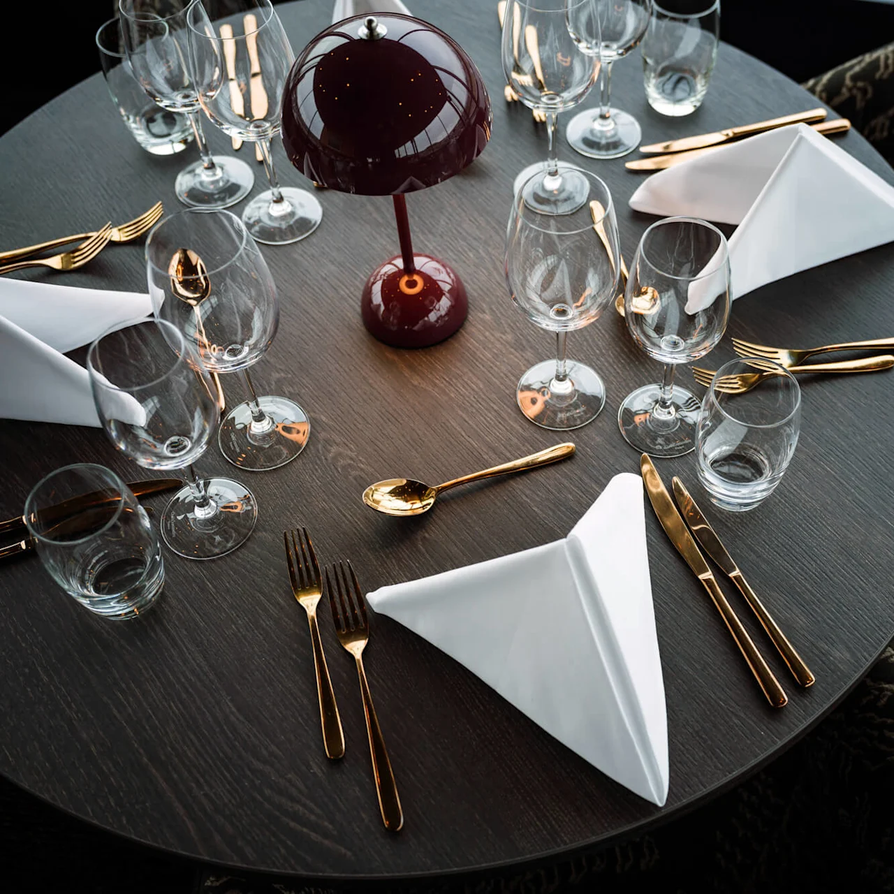 Round dining table set with folded white napkins, golden cutlery, multiple wine glasses, and a small red lamp in the center, surrounded by patterned chairs.