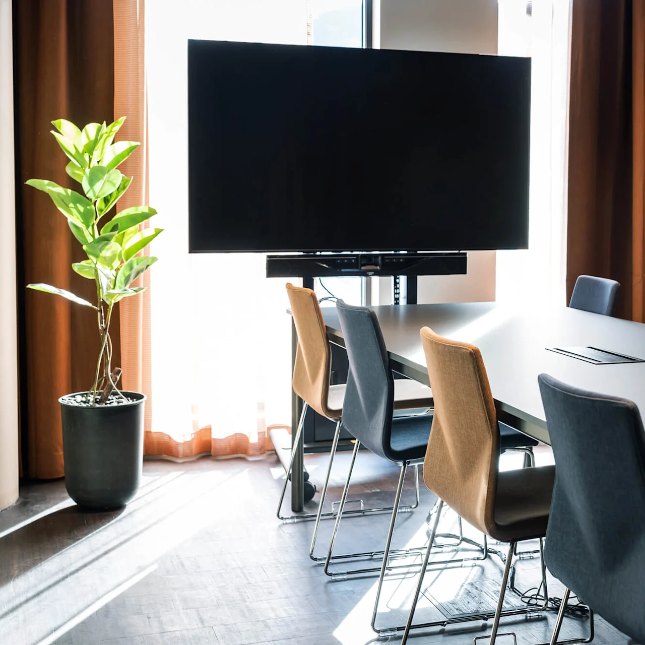 Konferencebord omringet af stole, placeret under en sort skærm, med en grøn potteplante ved siden af gulvlange gardiner, hvilket oplyser det moderne rum med naturligt lys.