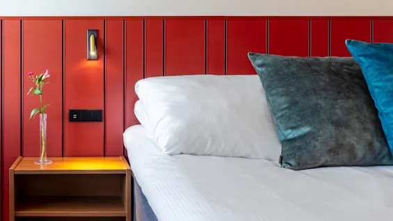 A bedroom corner with a white bed, blue and gray pillows, against a red paneled wall with a lamp and flower vase on a wooden nightstand.