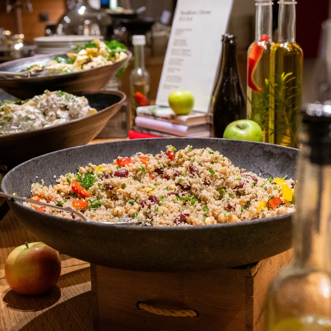 Colorful quinoa salad in a large bowl at a buffet with oils and apples in the background.