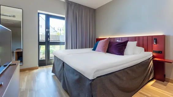 A neatly made bed in a modern hotel room with a balcony door open, showing an urban view.