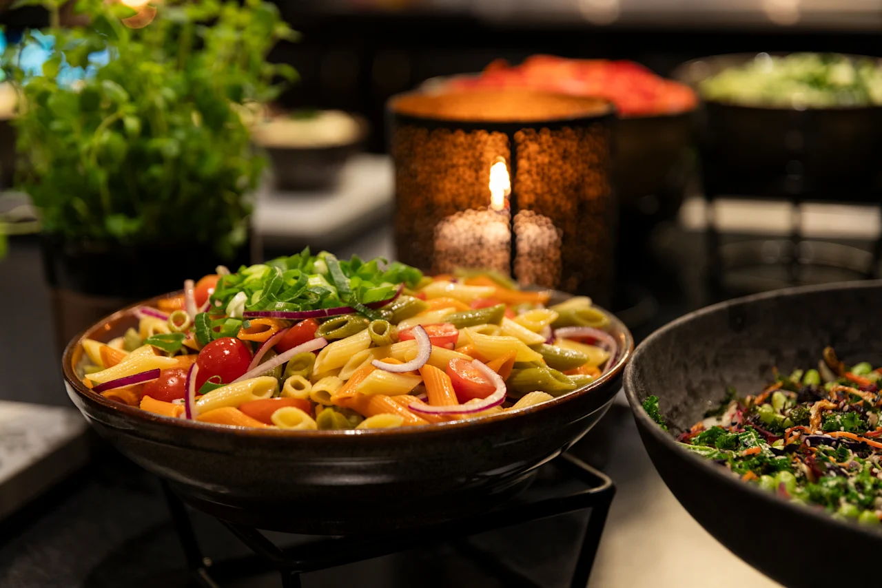 A vibrant pasta salad with tomatoes, onions, and greens on a buffet table at Home Hotel Grand Bodø, with a warm candle glow in the background.