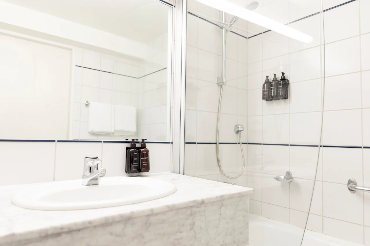 Valoisa ja puhdas kylpyhuone, jossa on valkoinen pesuallas, suuri peili ja suihku-amme-yhdistelmä seinään kiinnitetyillä annostelijoilla.