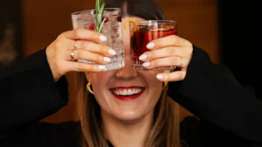 Henkilö, jolla on pitkät hiukset, pitää kahta cocktailia kasvonsa edessä, peittävät ne. Yhdessä lasissa on kirkasta nestettä ja rosmariinia; toisessa on punainen juoma. Tausta on sumea.