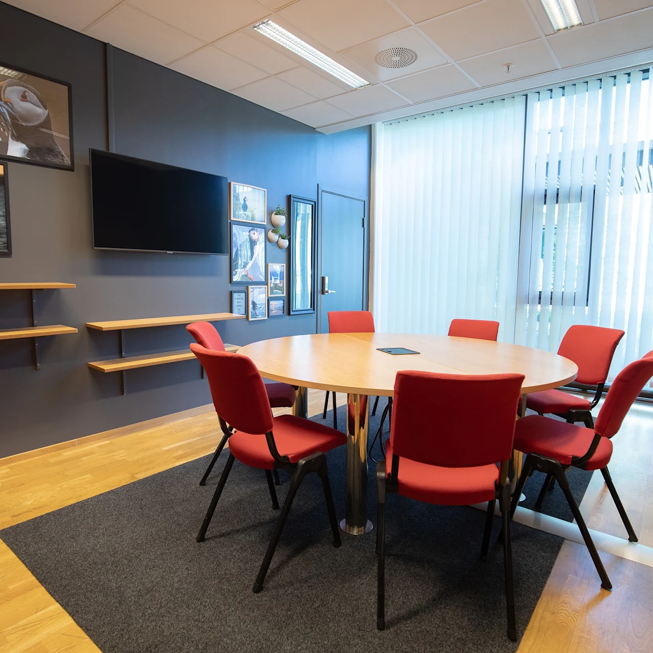 Et rundt bord omgitt av seks røde stoler står på et teppe i et konferanserom med en TV, veggkunst av fugler og gulv-til-tak-vinduer.