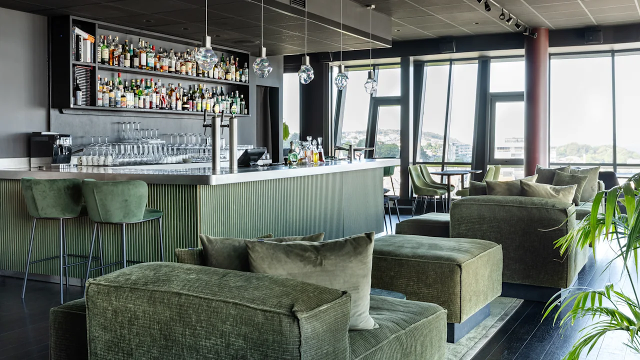 Luksuriøs bar med høje hylder fyldt med forskellige flasker, glas opstillet på skranken og bløde grønne siddepladser; store vinduer med panoramaudsigt.