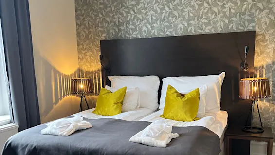 A neatly made bed with grey bedding and yellow accent pillows, flanked by lamps, in a room with patterned wallpaper.