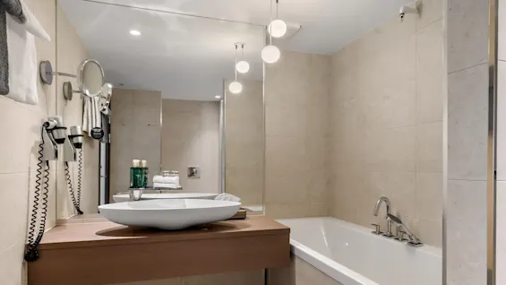 Et moderne badeværelse med en hvid håndvask og spejl på en træbænk, ved siden af et badekar. Håndklæder og toiletartikler er pænt arrangeret mod beige flisevægge.
