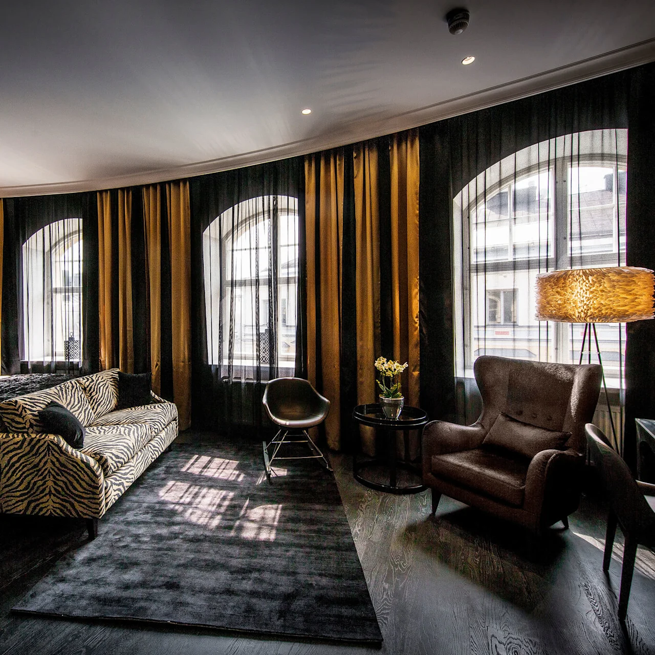 Et luksuriøst hotellrom har en stor seng med dekorative puter, en sebrastrøket sofa, elegant lenestol og høy lampe. Gulv-til-tak-vinduer er dekket av mørke gardiner.
