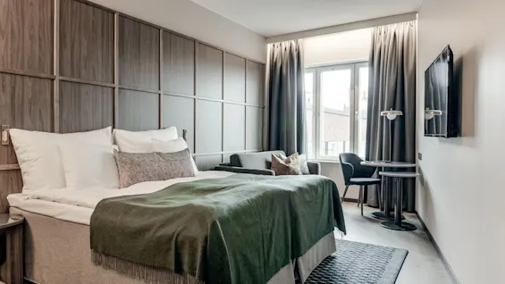 En pænt opredt seng med et grønt tæppe står mod en væg med træpanel i et hotelværelse, ved siden af en lille siddeområde og et vindue med mørke gardiner.
