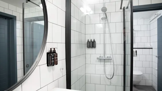Rundt spejl reflekterer en moderne badeværelsesscene med glasomsluttet bruser, sorte armaturer og hvide fliser. Et toilet er synligt i baggrunden.