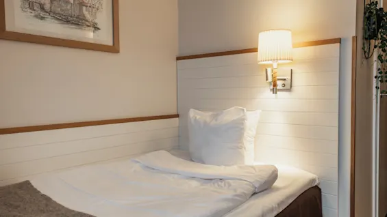En mysig enkelsäng med vita lakan och en varm vägglampa, som inbjuder till en vilsam vistelse i ett bekvämt rum.