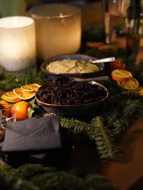Ett bord håller skålar med torkad frukt och dipp omgivna av tallkvistar och citrusskivor, nära tända ljus och en dryckesdispenser i en mysig, svagt upplyst miljö.