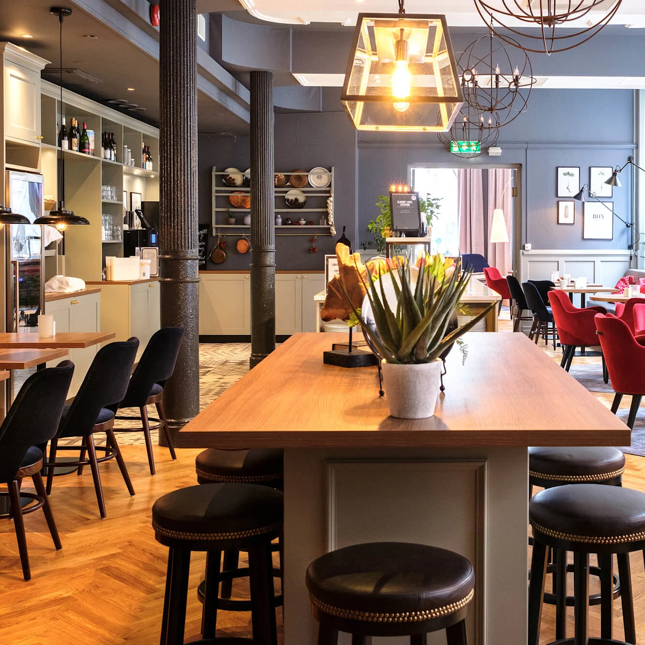 A cozy restaurant interior with red chairs, wooden tables, and stylish lighting fixtures. Elegant bar stools align with a counter.