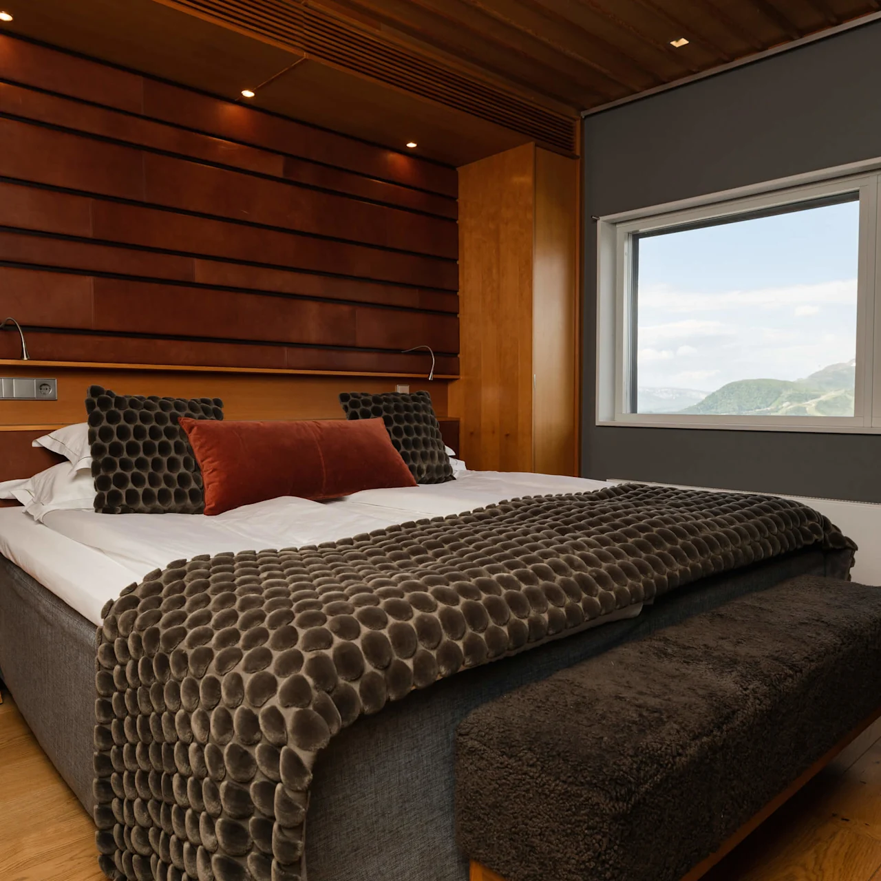 A neatly made bed with decorative pillows in a room with wooden accents and a mountain view through the window.