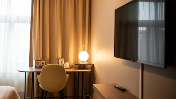 Ett skrivbord med en rund lampa och en radio står vid ett fönster med beige gardiner. En stol står vid skrivbordet. En TV är monterad på väggen.