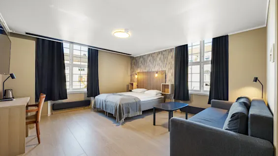 Lyst og romslig hotellrom med dobbeltseng, sofa, skrivebord og store vinduer.