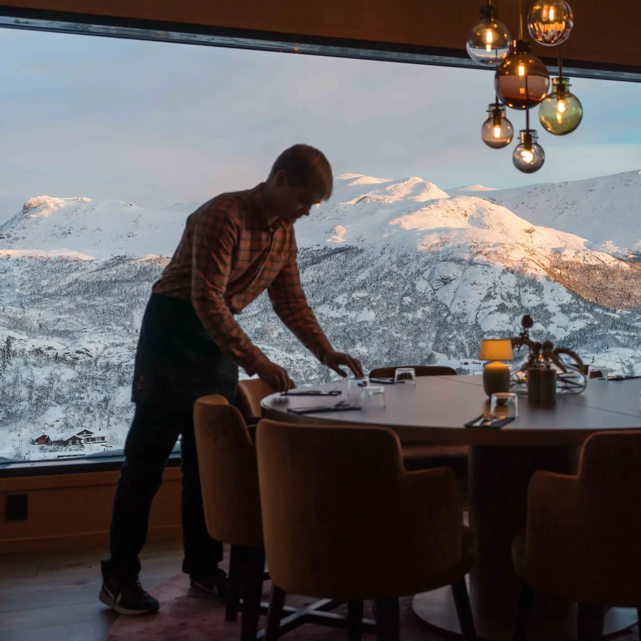 A waiter in a plaid shirt sets a dining table inside a cozy restaurant, framed by large windows showing snowy mountains during sunset. Warm hanging lights illuminate the scene.