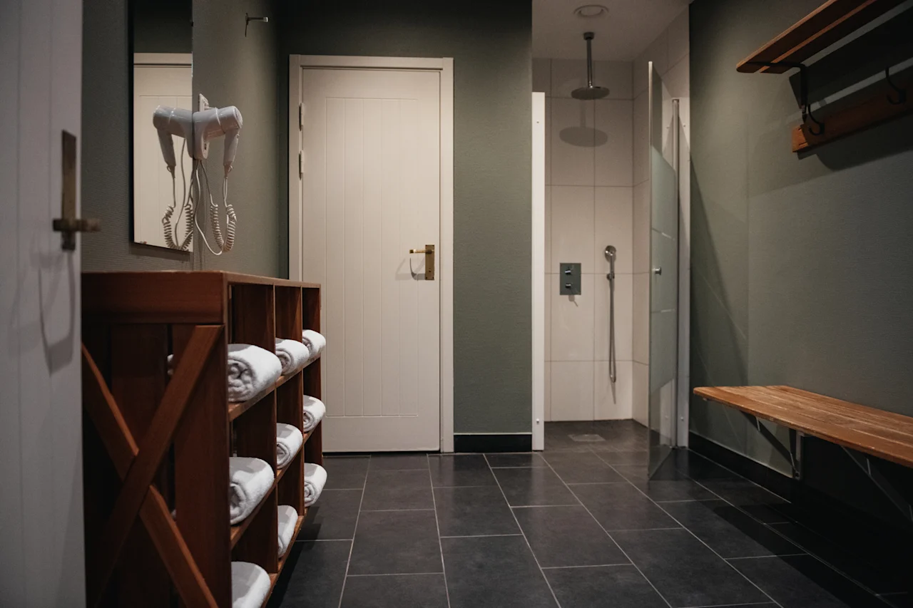 Ett rent och modernt omklädningsrum med duschar, en träbänk och fräscha handdukar. En hårtork hänger vid en spegel.