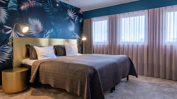 A bed with a dark quilt and white pillows sits in a room with a vibrant leaf-patterned accent wall and curtains covering large windows. Two lamps flank the bed.