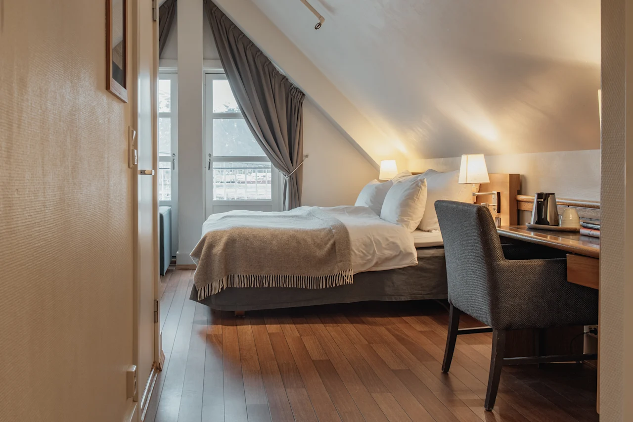 A cozy hotel room with a comfortable bed, a desk, and a chair, featuring a sloped ceiling and a window with curtains.
