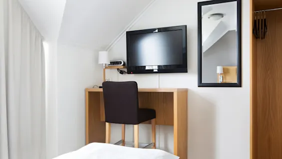 TV festet på veggen over et trebord med en svart stol i et lite hotellrom. Et speil er på veggen ved siden av et garderobeskap.