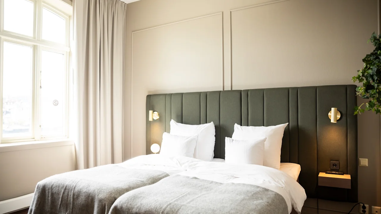 Et lyst hotellrom med to komfortable senger, en mørkegrønn sengegavl og et stort vindu med lyse gardiner.