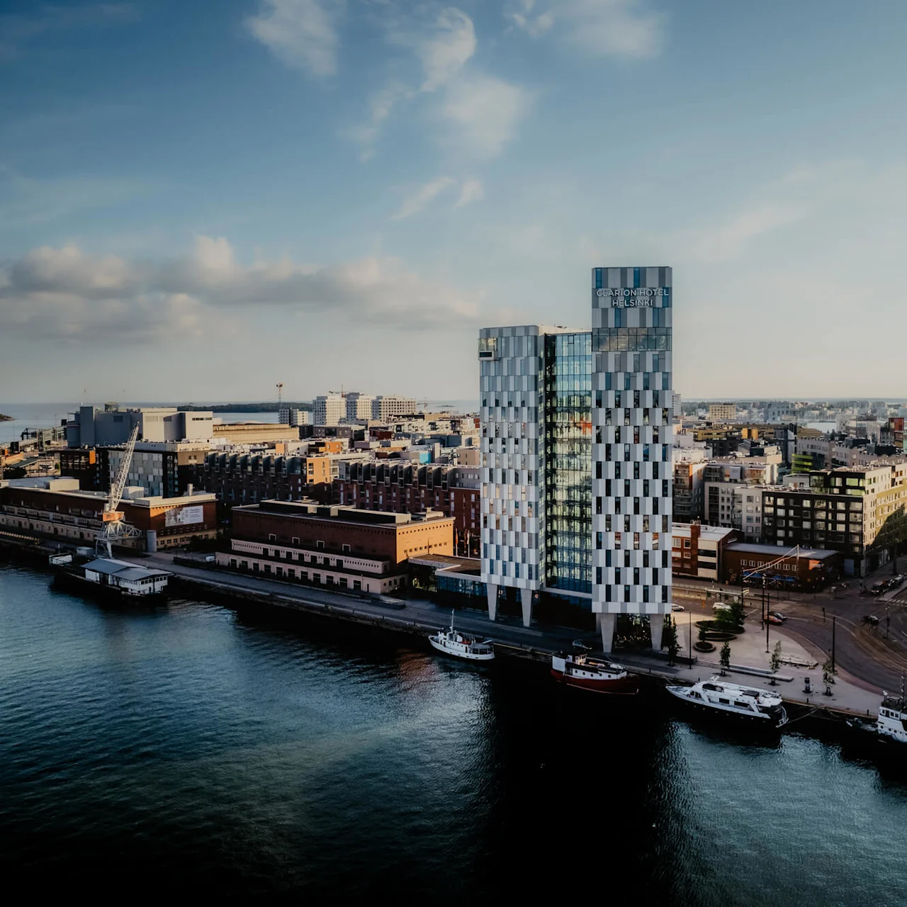 En mønstret skyskraper med teksten "Clarion Hotel Helsinki" står ved vannkanten og er omgitt av andre bystrukturer. Båter fortøyd langs vannet under en delvis skyet himmel.