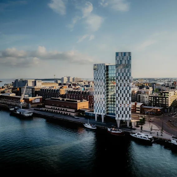 Kuvioitu korkea rakennus, jossa lukee "Clarion Hotel Helsinki", seisoo veden äärellä, ympäröitynä muilla kaupunginrakennuksilla. Venelaitureita veden äärellä puolipilvisen taivaan alla.