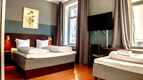 Ett mysigt hotellrum med två sängar, en väggmonterad TV och ett fönster med naturligt ljus. Rummet har en varm och inbjudande atmosfär.