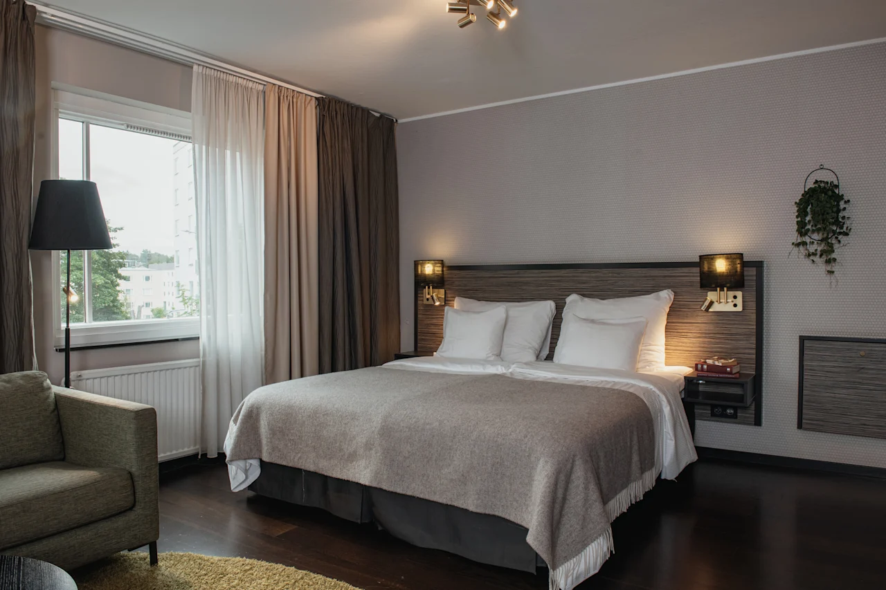 A cozy hotel room with a large bed, a comfortable armchair by the window, and warm lighting, inviting relaxation.