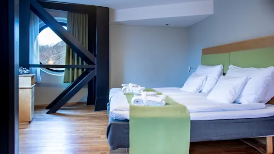 A neatly made bed with white linens and a green throw sits in a wooden-floored room, near a circular window framed with black beams.