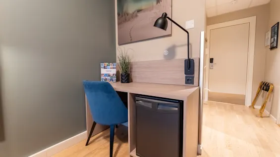 Et koselig skrivebordsområde på et hotellrom med en blå stol, lampe og minikjøleskap. En brosjyre med tittelen TROMSO ligger på skrivebordet.