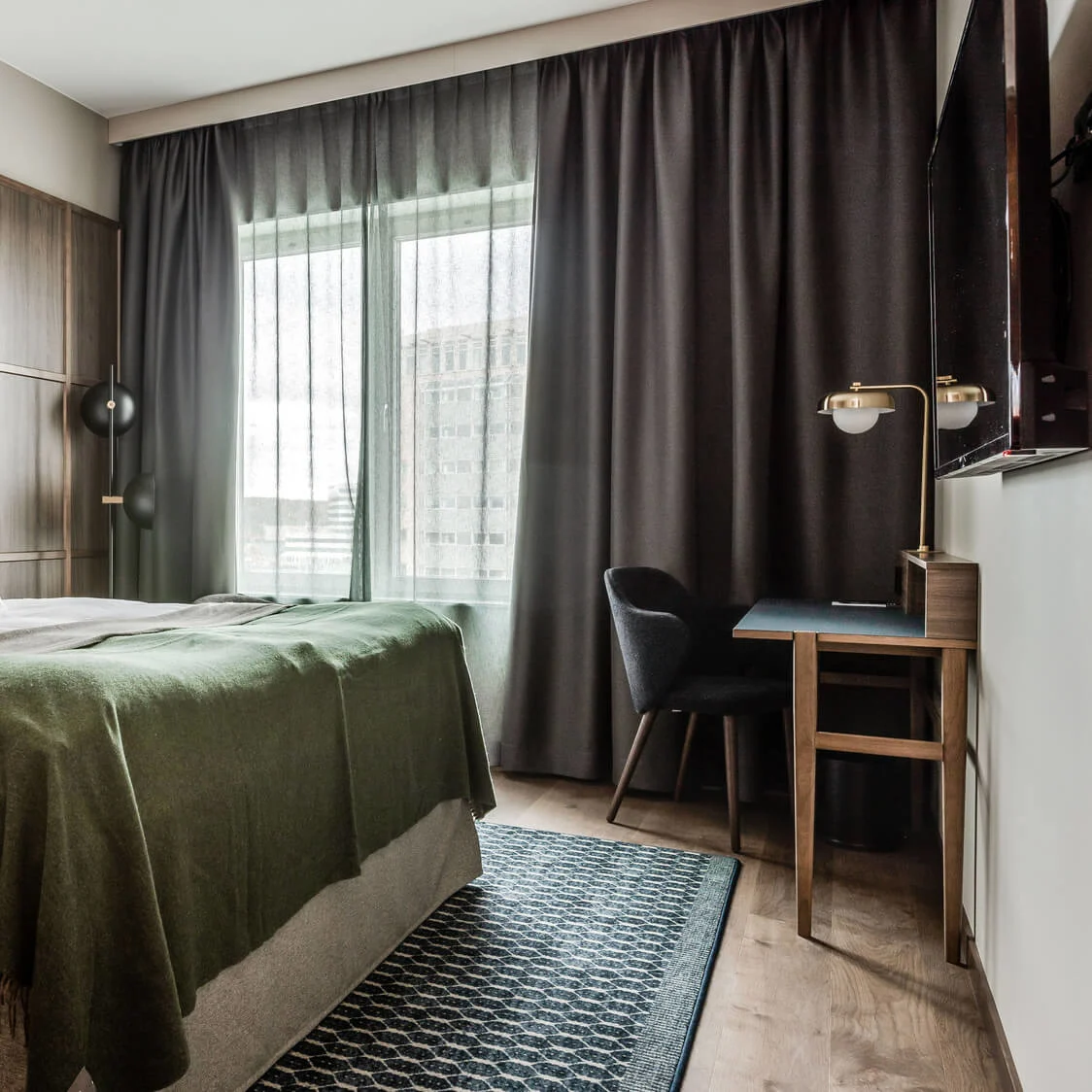 En seng med hvide puder og et grønt tæppe står mod en træpanelvæg. Et skrivebord med stol og lampe er ved siden af, med gardiner og et vindue i baggrunden.