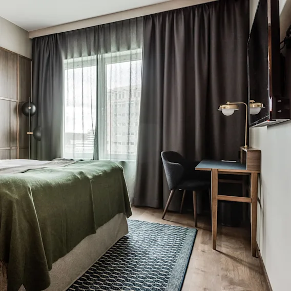 En säng med vita kuddar och en grön filt står mot en träpanelvägg. Ett skrivbord med stol och lampa är bredvid, med gardiner och ett fönster i bakgrunden.