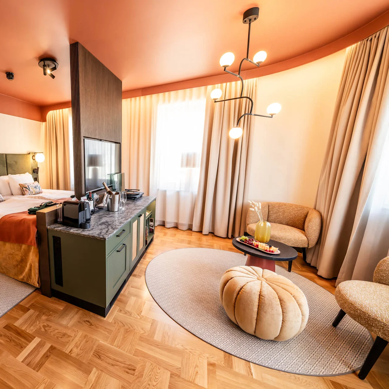 A stylish hotel room features a neatly made bed, a dividing unit with a TV, cozy seating area with a table, soft lighting, and decorative accents in warm, earthy tones.