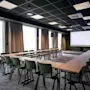 A modern conference room with tables and chairs arranged in a U-shape, facing a large projection screen. Large windows line one wall.