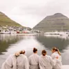 Personer i badekåber sidder ved vandet og ser ud over en havn og bjerge