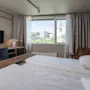 En seng er pænt redt med hvidt sengetøj på et hotelværelse. Et skrivebord med lampe og TV står i nærheden. Et vindue viser udsigt over en by med et kirketårn.