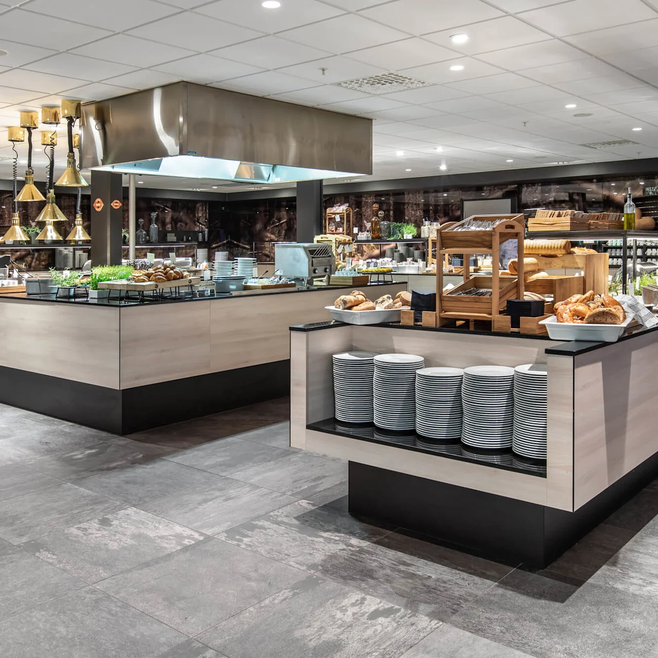 Buffet setup displaying assorted breads, plates, and condiments, arranged on modern counters in a well-lit dining area, featuring decorative lighting and a visible exhaust hood.