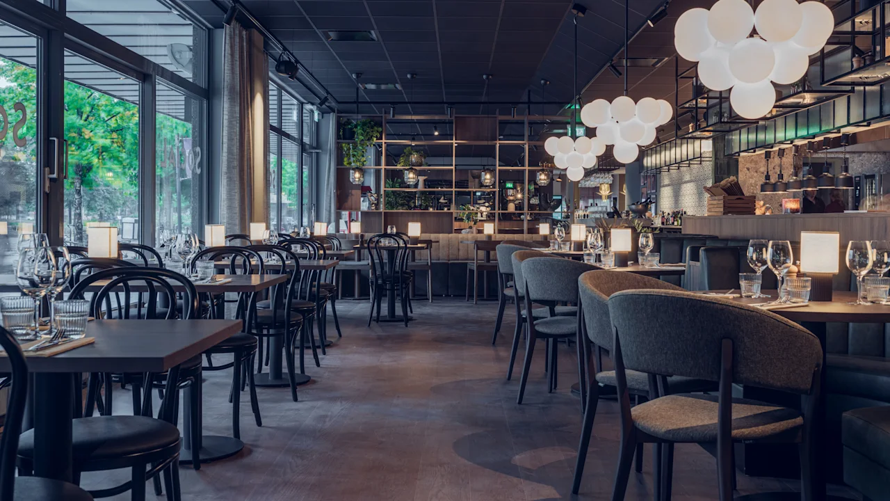 Matbord och stolar står ordentligt i en modern, stilren restaurang, upplyst av sfäriska taklampor. Stora fönster släpper in naturligt ljus och ger utsikt över grönska utanför. Ingen synlig text.