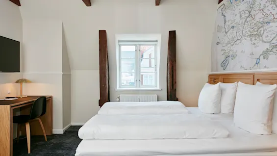 Et hyggeligt hotelværelse med to senge, et skrivebord og et stort kort på væggen, med synlige træbjælker og et lyst vindue.