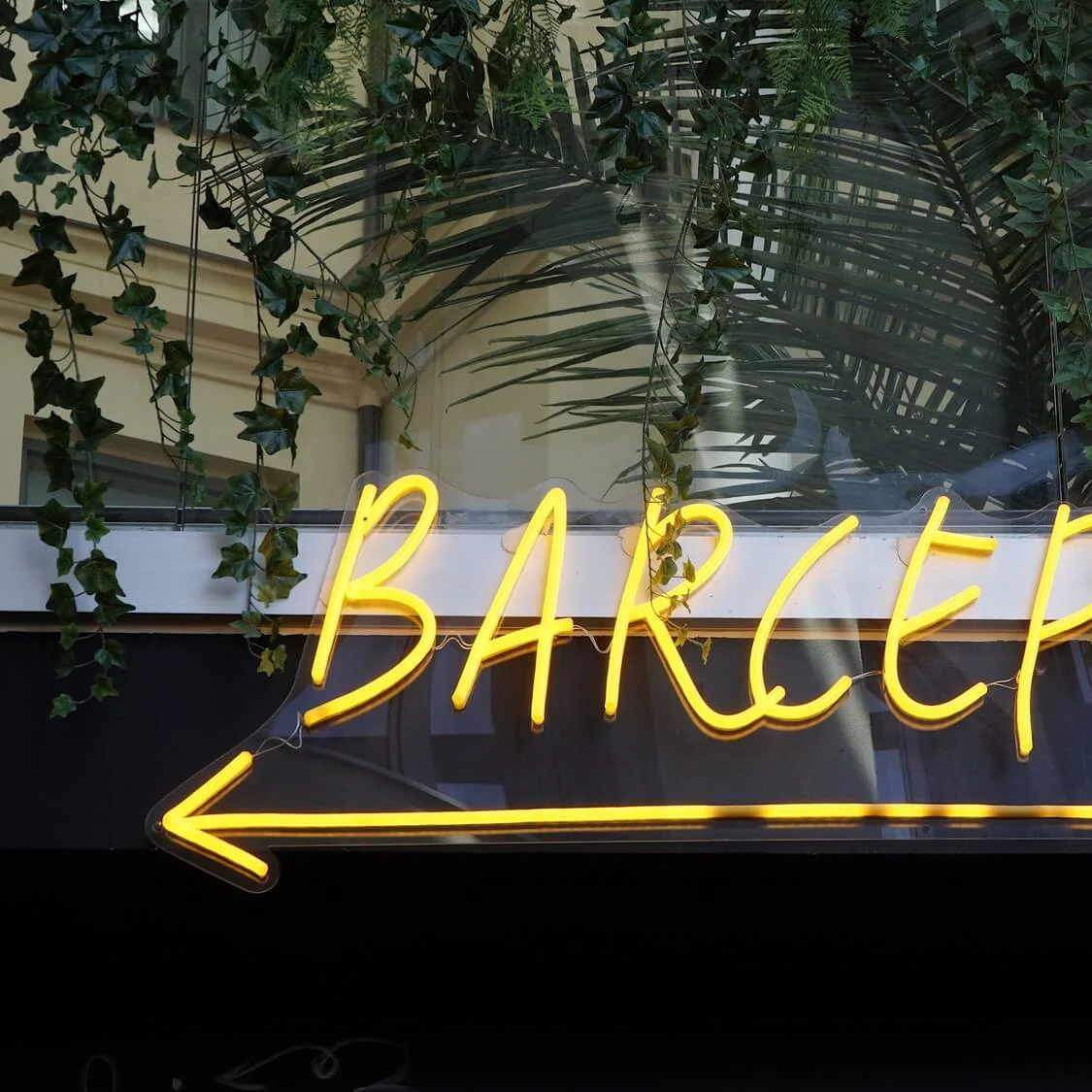Neon sign spelling "BARCEPTION" with an arrow, against a backdrop of plants and window reflections.