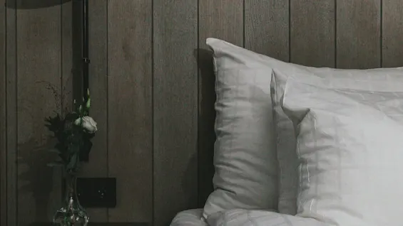 Bed with white pillows and duvet rests against a wooden paneled wall; a bedside table holds a vase with flowers and a copper lamp.