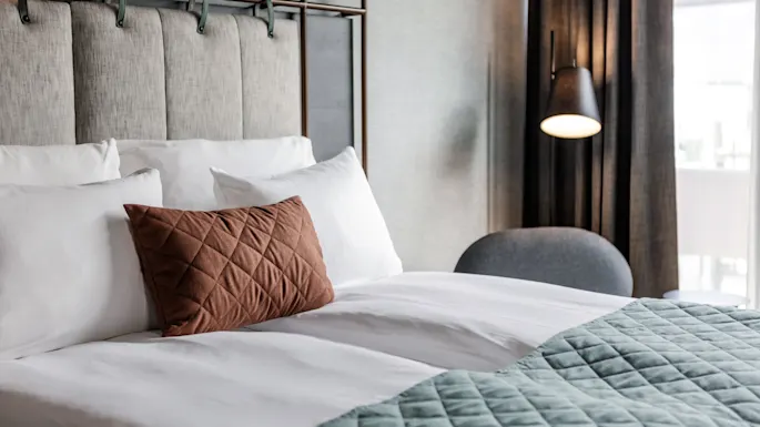 A neatly made bed with white pillows and a brown, quilted cushion. A green quilted blanket covers part of the bed. A soft light lamp hangs beside gray curtains.