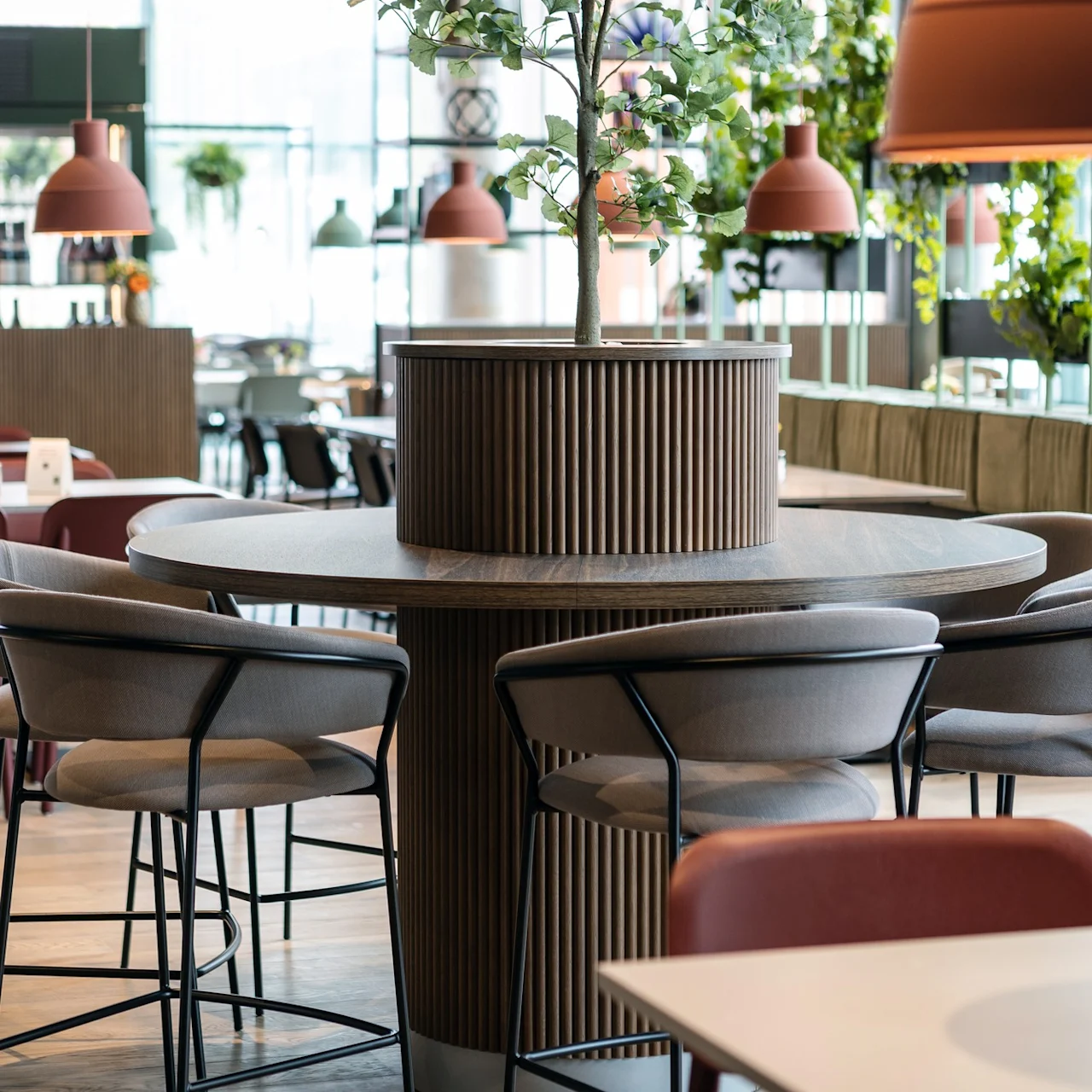 Runda träbord omger en central växt, med stoppade stolar. Hängande lampor och glasfönster lyser upp den moderna, mysiga restaurangens interiör med gröna växter.