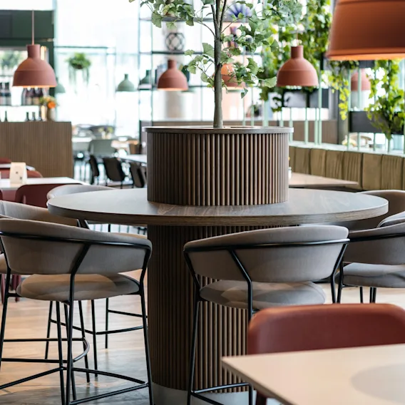 Runde treborde omgiver en central plante, med polstrede stole. Hængende lamper og glasvinduer lyser op i den moderne, hyggelige restaurantindretning med grønne planter.